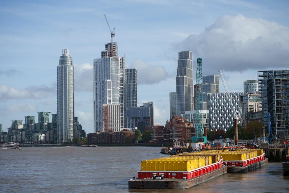 A panoramic view of a modern cityscape with a river in the foreground, featuring a stone and metal bridge spanning across the water. Behind the bridge, there are numerous high-rise buildings with vari